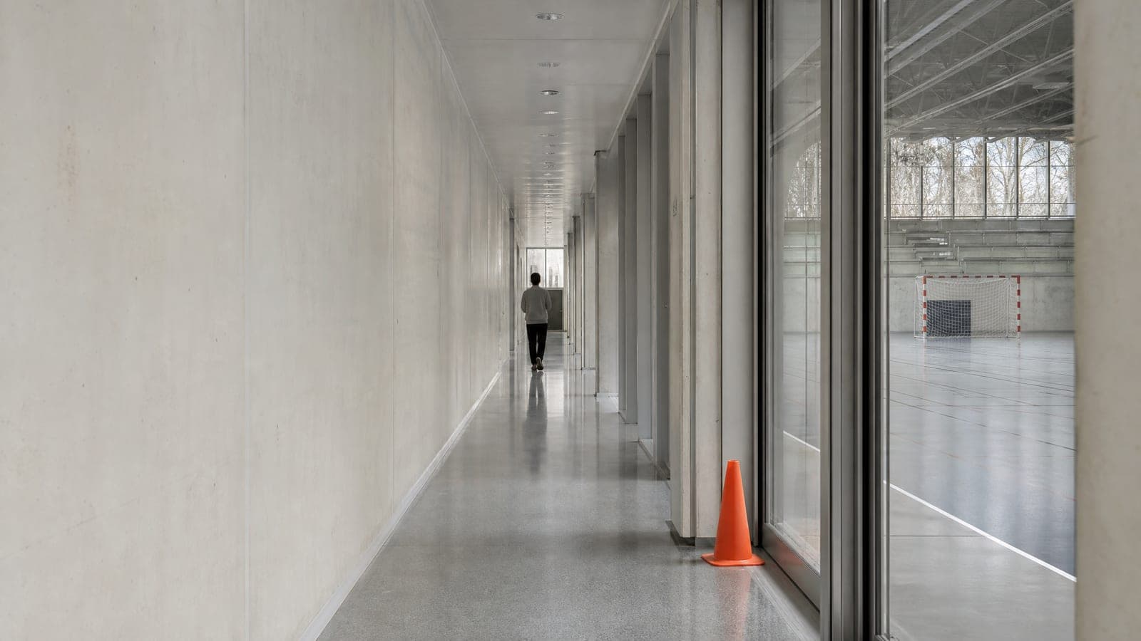 A long pale sports-hall corridor seen down its length with one distant figure walking away in a plain sweatshirt.
