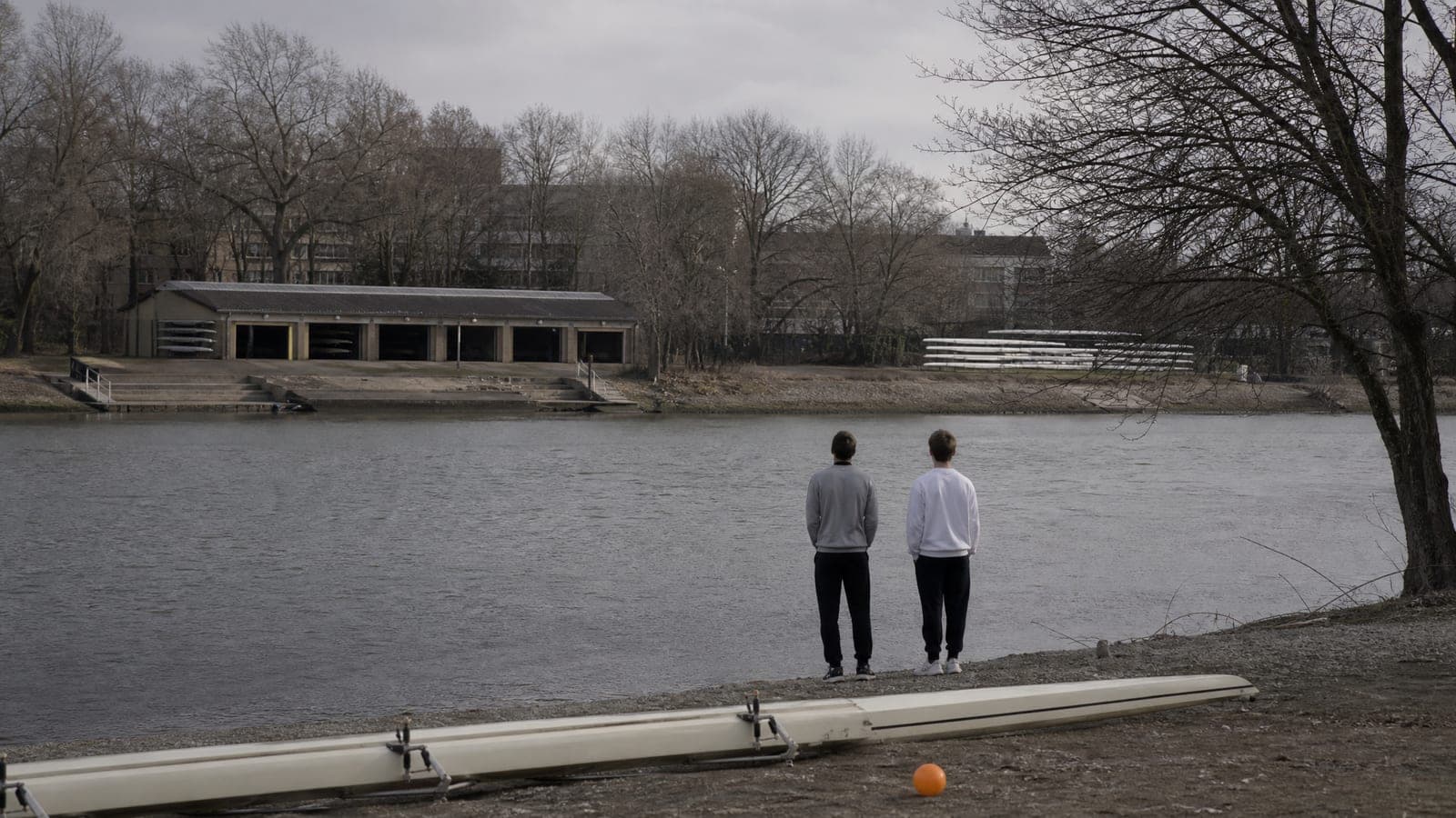 An overcast river bank with a boathouse in the middle distance and two figures at the water's edge seen from behind in plain sweatshirts.